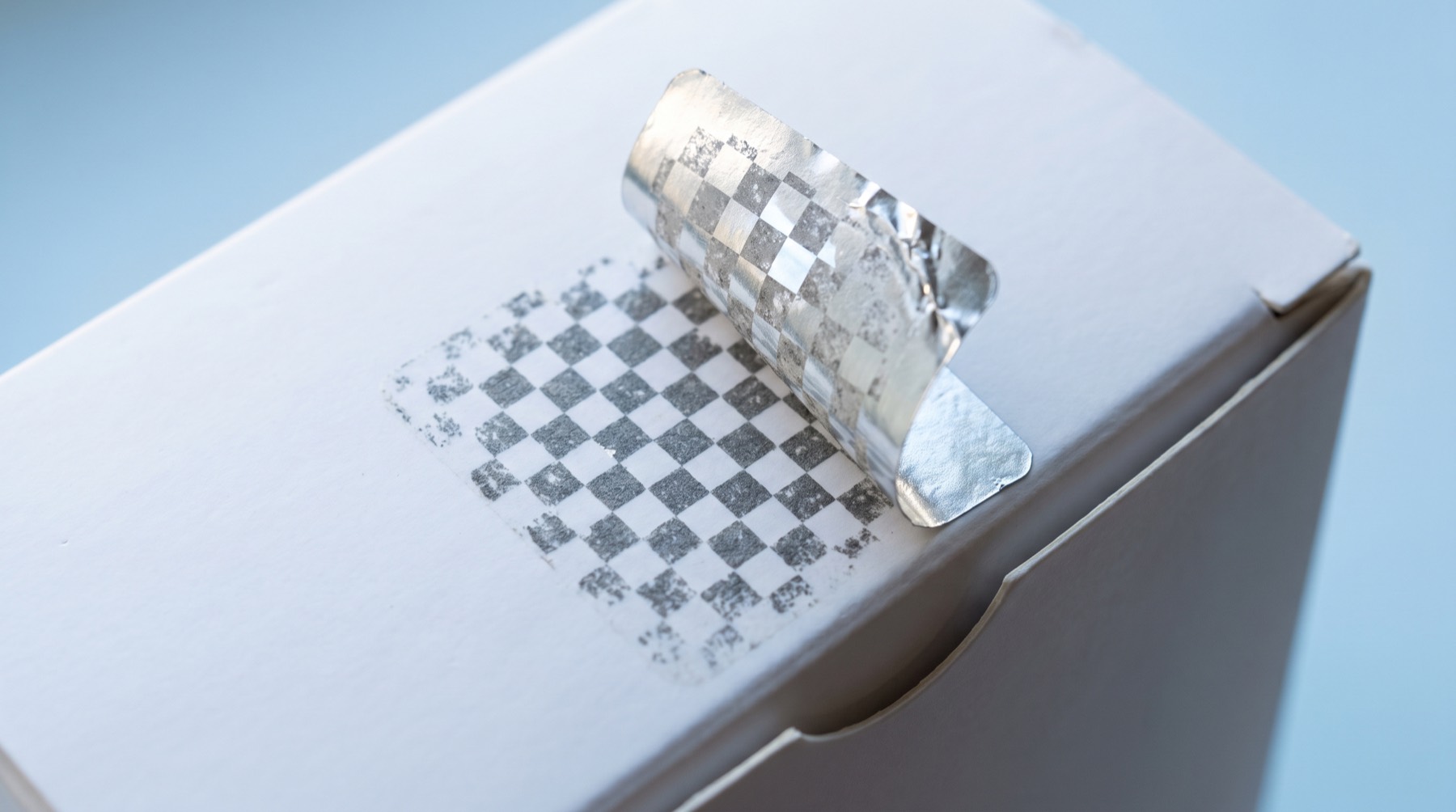 A metallic security label half peeled from a white box, leaving a checkerboard residue on the surface